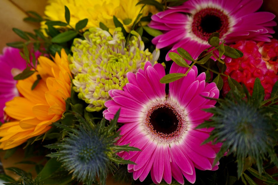 A vibrant floral arrangement featuring large pink gerbera daisies with dark centers, complemented by bright yellow sunflowers with orange accents, green chrysanthemum blooms with tightly packed petals