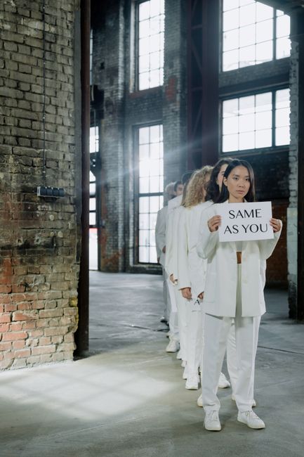 A line of women dressed in white suits standing inside an industrial-style building with large grid windows and exposed brick walls. The woman at the front holds a white sign with black text reading '