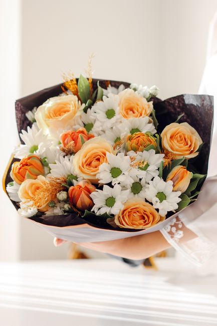 A bouquet of fresh flowers held by a person in a white sleeve, featuring large peach roses with soft, velvety petals, white daisies with yellow centers, and small orange ranunculus blooms. The arrange