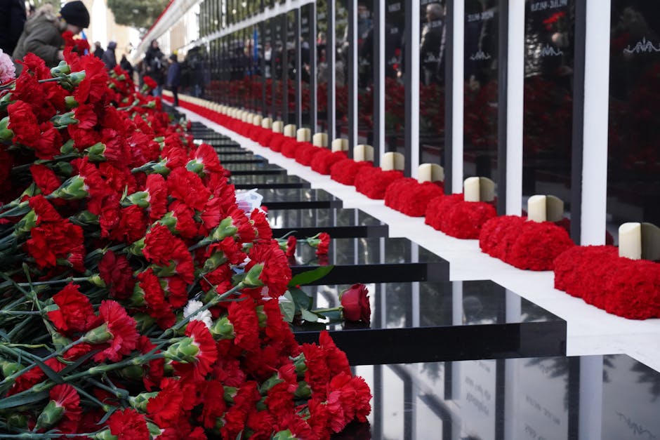A line of vibrant red carnations, fresh and with lush green stems, arranged on a memorial platform with black polished stone surfaces reflecting the flowers. Behind the carnations, a white fence with