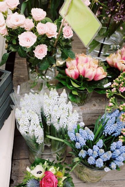 A vibrant display of fresh floral arrangements featuring a bouquet of pale pink roses with lush green foliage, placed in a clear glass vase on a rustic wooden surface. Surrounding it are more flowers